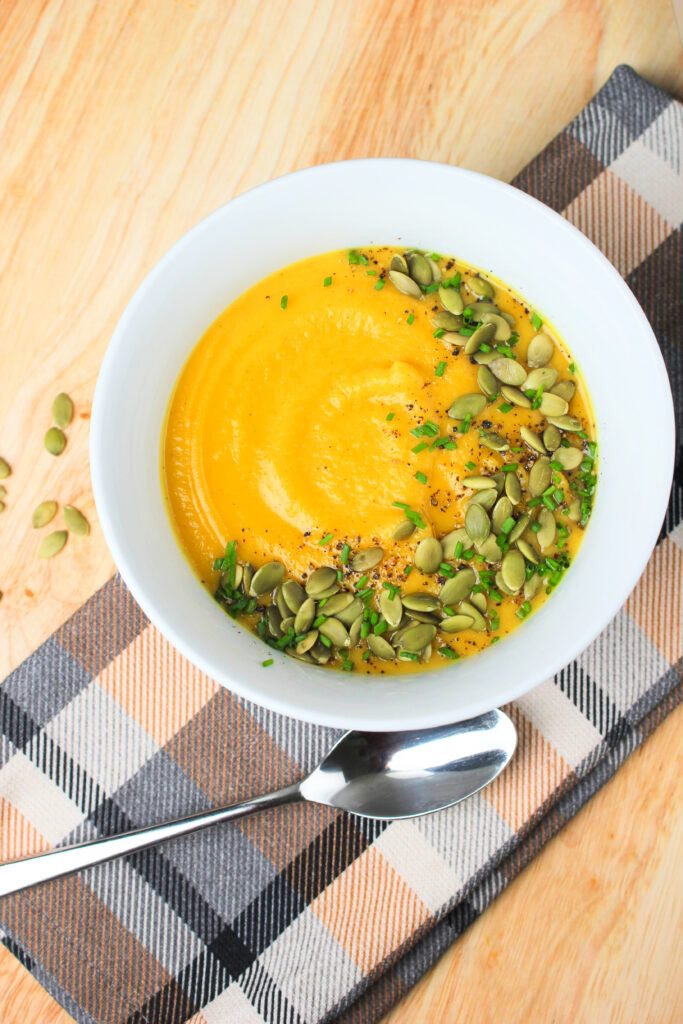 The pumpkin soup is in a white soup bowl and garnished with pumpkin seeds, chives, and pepper.