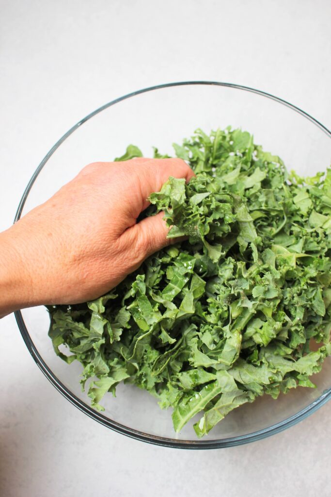 The kale is in a large clear bowl. I am massaging olive oil into the kale with my hands.