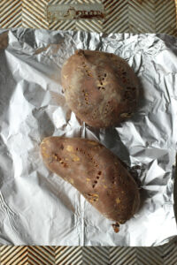 Two sweet potatoes are poked with a fork and placed on a baking sheet and ready to be roasted.