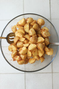 The seasonings have been tossed into the chopped potatoes in a clear glass bowl.